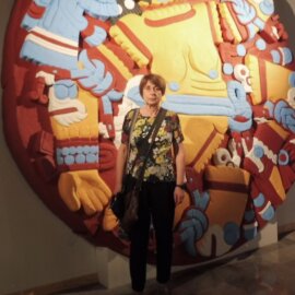 Daniele Dehouve stands indoors in front of a large, colorful circular artwork inspired by Mesoamerican design. The mural features bold patterns and stylized figures in red, yellow, blue, and white, arranged in a radial composition.