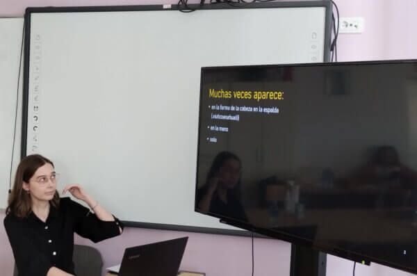 Gabriela Piszczatowska prowadzi prezentację przy biurku z laptopem Lenovo; za nią widoczny jest baner z tekstem w języku hiszpańskim dotyczącym motywu „xuihcoanahualli”.