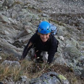 Katarzyna Szoblik is rock climbing outdoors on rugged terrain, wearing a blue helmet and a backpack. She is ascending a steep, rocky slope using both hands and feet.