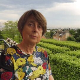 Daniele Dehouve está de pie en una terraza al aire libre, con una vista panorámica de París al fondo.