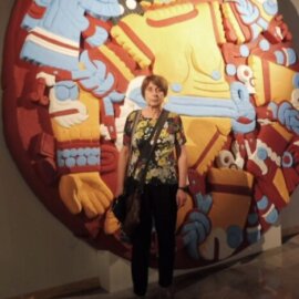 Daniele Dehouve stoi wewnątrz budynku przed dużym, kolorowym, okrągłym dziełem sztuki inspirowanym wzornictwem mezoamerykańskim. Mural przedstawia wyraziste wzory i stylizowane postacie w kolorach czerwonym, żółtym, niebieskim i białym, ułożone w kompozycji radialnej.