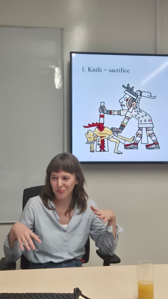 Julia Madajczak siedzi przy biurku w sali wykładowej, a za nią wyświetlany jest slajd z grafiką rytualnej ofiary z kodeksu nahua i podpisem „1. Knife = sacrifice”.