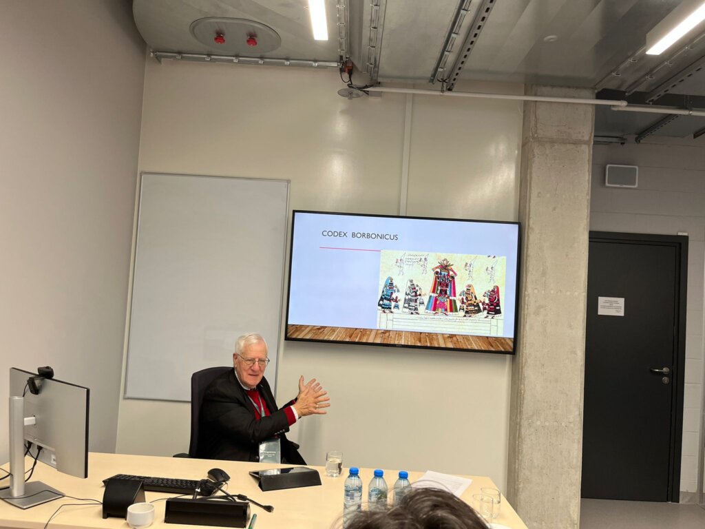 John F. Schwaller siedzi przy biurku w sali konferencyjnej, a na ekranie wyświetla się slajd zatytułowany „CODEX BORBÓNICUS” z ilustracją kodeksu.