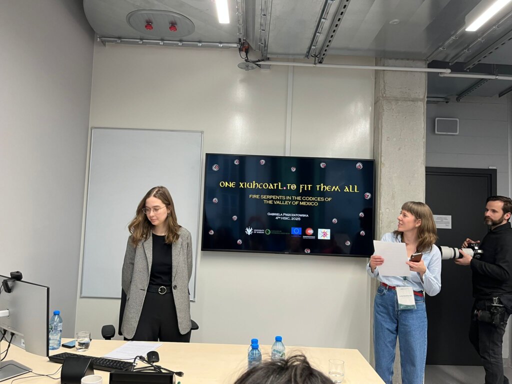 Gabriela Piszczatowska stoi przy ekranie, na którym wyświetla się tytuł referatu “ONE XIUHCOATL TO FIT THEM ALL: Fire Serpents in the Codices of the Valley of Mexico”, Julia Madajczak dokonuje wprowadzenia, a fotograf dokumentuje wydarzenie.