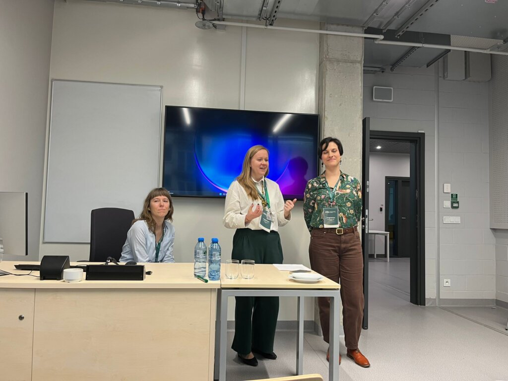 Julia Madajczak siedzi przy biurku, a Agnieszka Brylak i Justyna Kowalczyk-Kądziela stoją obok niej w nowoczesnej sali konferencyjnej.