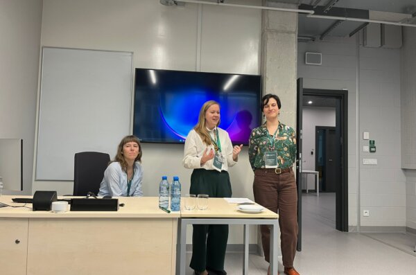 Julia Madajczak siedzi przy biurku, a Agnieszka Brylak i Justyna Kowalczyk-Kądziela stoją obok niej w nowoczesnej sali konferencyjnej.