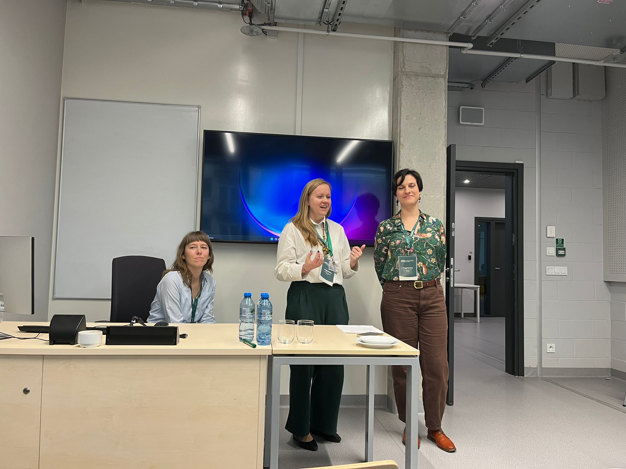 Julia Madajczak siedzi przy biurku, a Agnieszka Brylak i Justyna Kowalczyk-Kądziela stoją obok niej w nowoczesnej sali konferencyjnej.