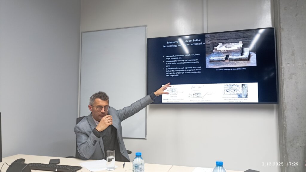 Jarosław Źrałka siedzi przy biurku w sali konferencyjnej i wskazuje na slajd o kąpielach parowych w Mezoameryce, zawierający tekst, zdjęcie łaźni z Joya de Cerén oraz diagramy struktur temazcal.