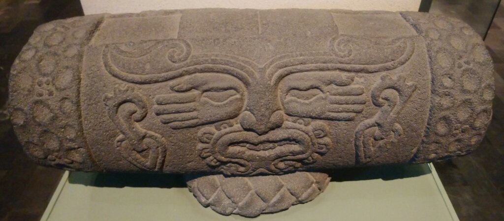 Tambor de piedra con un rostro tallado y motivos simétricos de manos.