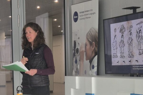 Dr. Molly H. Bassett stands at the front of a modern classroom, presenting beside a screen showing a historical illustration and a banner from the University of Warsaw.