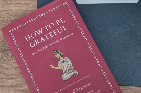 Un libro rojo titulado How to Be Grateful: An Aztec Guide to the Art of Gratitude está colocado sobre una superficie de madera junto a una carpeta azul oscuro de la Association of Nahuatl Scholars. La portada muestra una ilustración de una anciana noble.