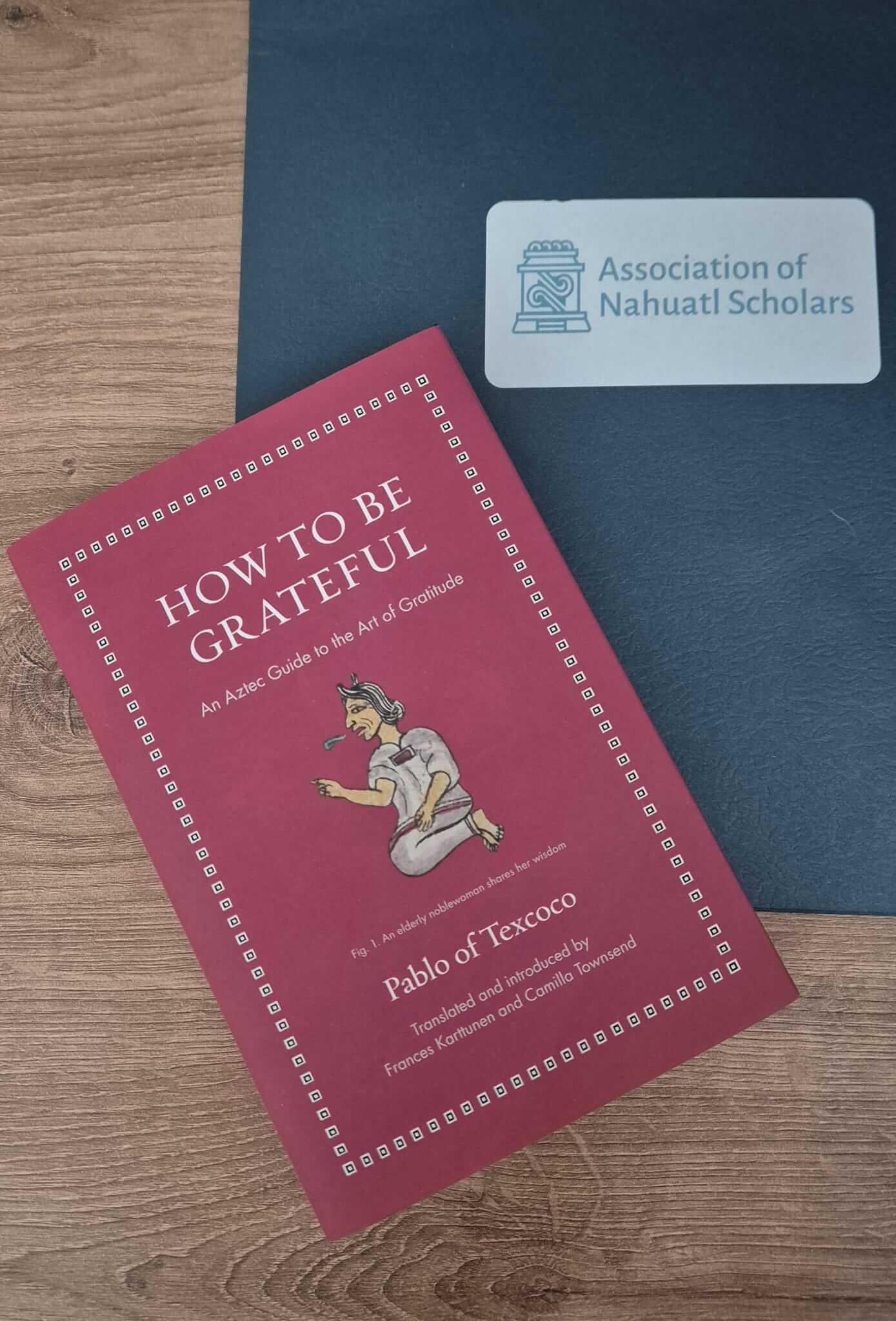 Un libro rojo titulado How to Be Grateful: An Aztec Guide to the Art of Gratitude está colocado sobre una superficie de madera junto a una carpeta azul oscuro de la Association of Nahuatl Scholars. La portada muestra una ilustración de una anciana noble.