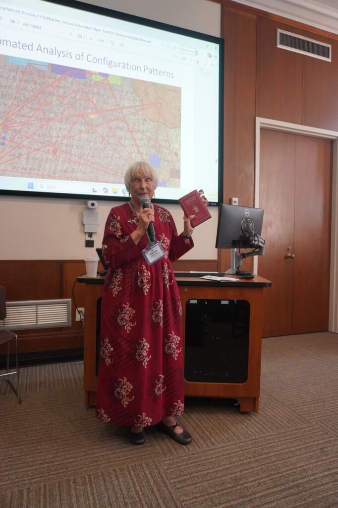 Una persona con un vestido rojo presenta junto a un atril, sosteniendo un micrófono y un libro. Detrás se ve una diapositiva titulada “Automated Analysis of Configuration Patterns”.