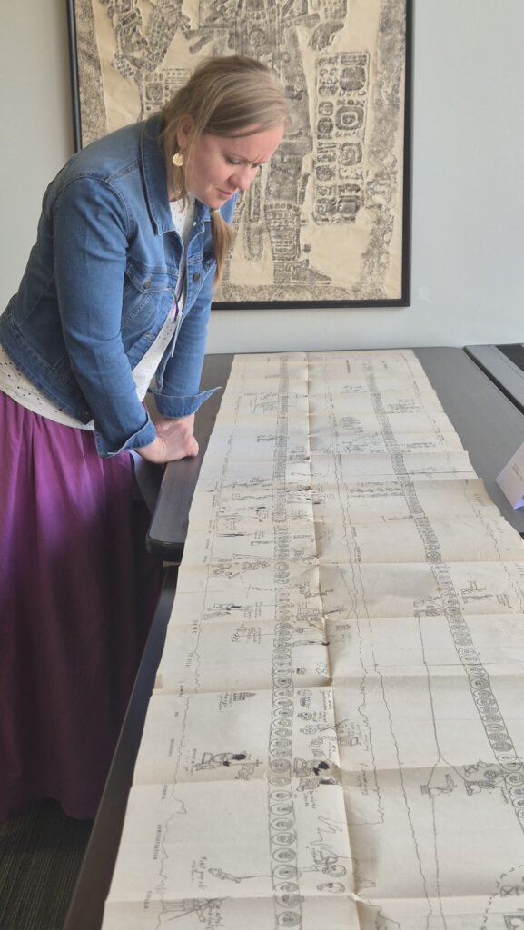 Agnieszka Brylak leans over a long unfolded manuscript filled with drawings and glyphs laid out on a table.