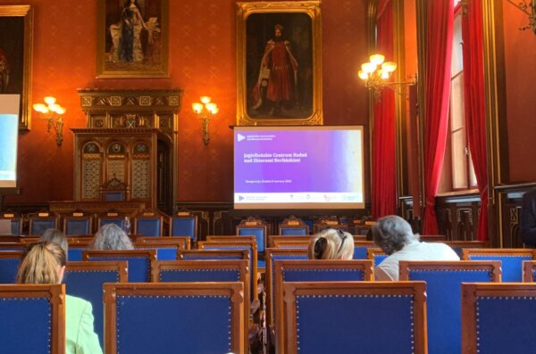 A lecture hall with red walls and portraits, where attendees sit facing a screen displaying a symposium presentation in Kraków.