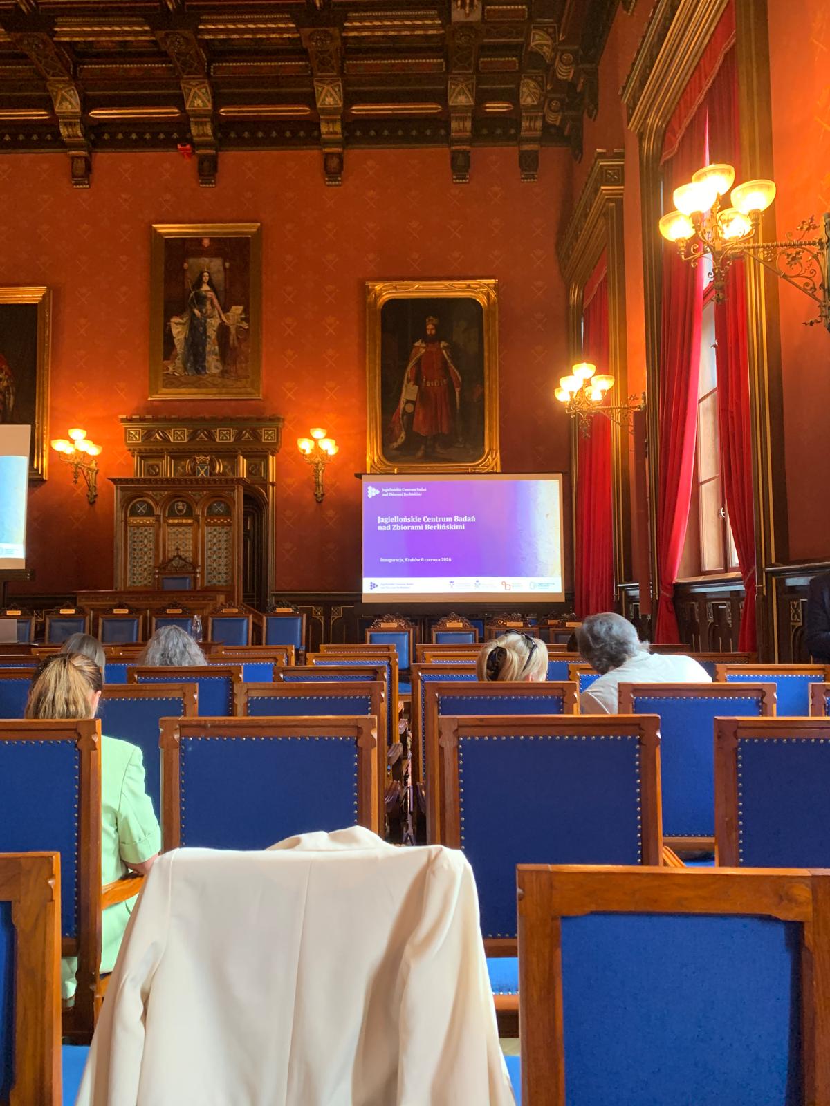 A lecture hall with red walls and portraits, where attendees sit facing a screen displaying a symposium presentation in Kraków.