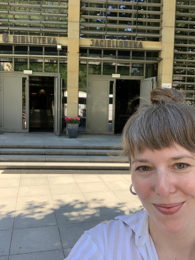 La doctora Julia Madajczak está de pie frente a la entrada de la Biblioteca Jaguelónica, con una fachada moderna de vidrio detrás.